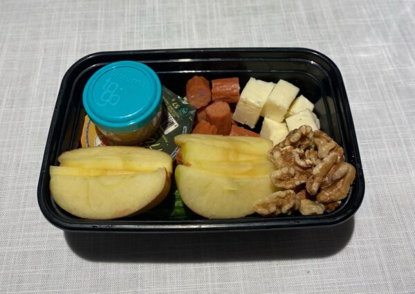 Lunch box with hummus in a container, peanut butter in a container, pepperoni and cheese cubes, walnuts, sliced apples, and a mini cucumber