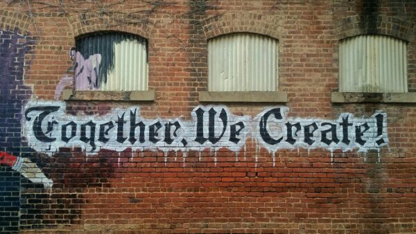 A brick wall with "Together, We Create!" spray painted on the side