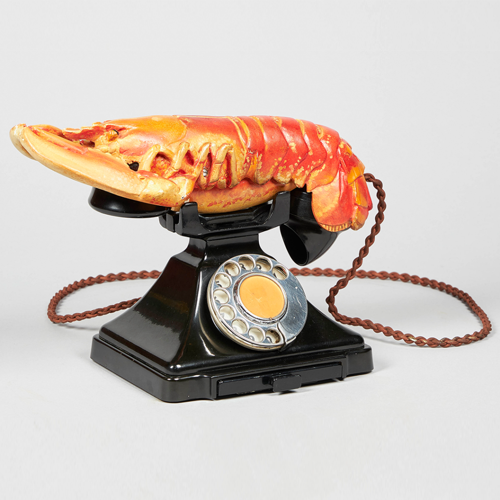 A rotary phone with a cooked lobster placed where the telephone should be. The phone is connected to the lobster by a chain.